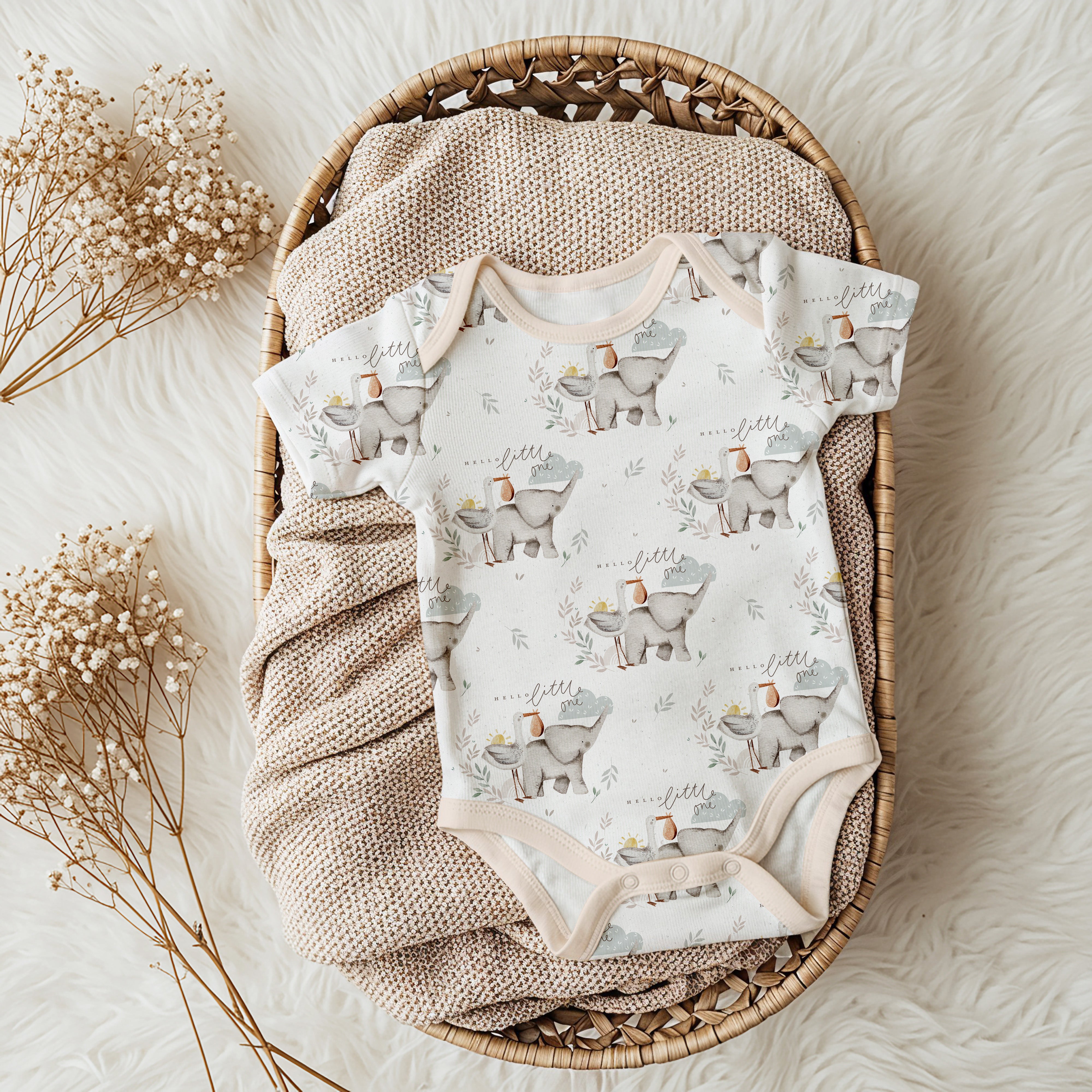 A baby short-sleeve bodysuit with a repeating elephant print, laid flat on a textured beige blanket inside a woven oval basket, with dried baby's breath flowers on a white faux-fur surface.