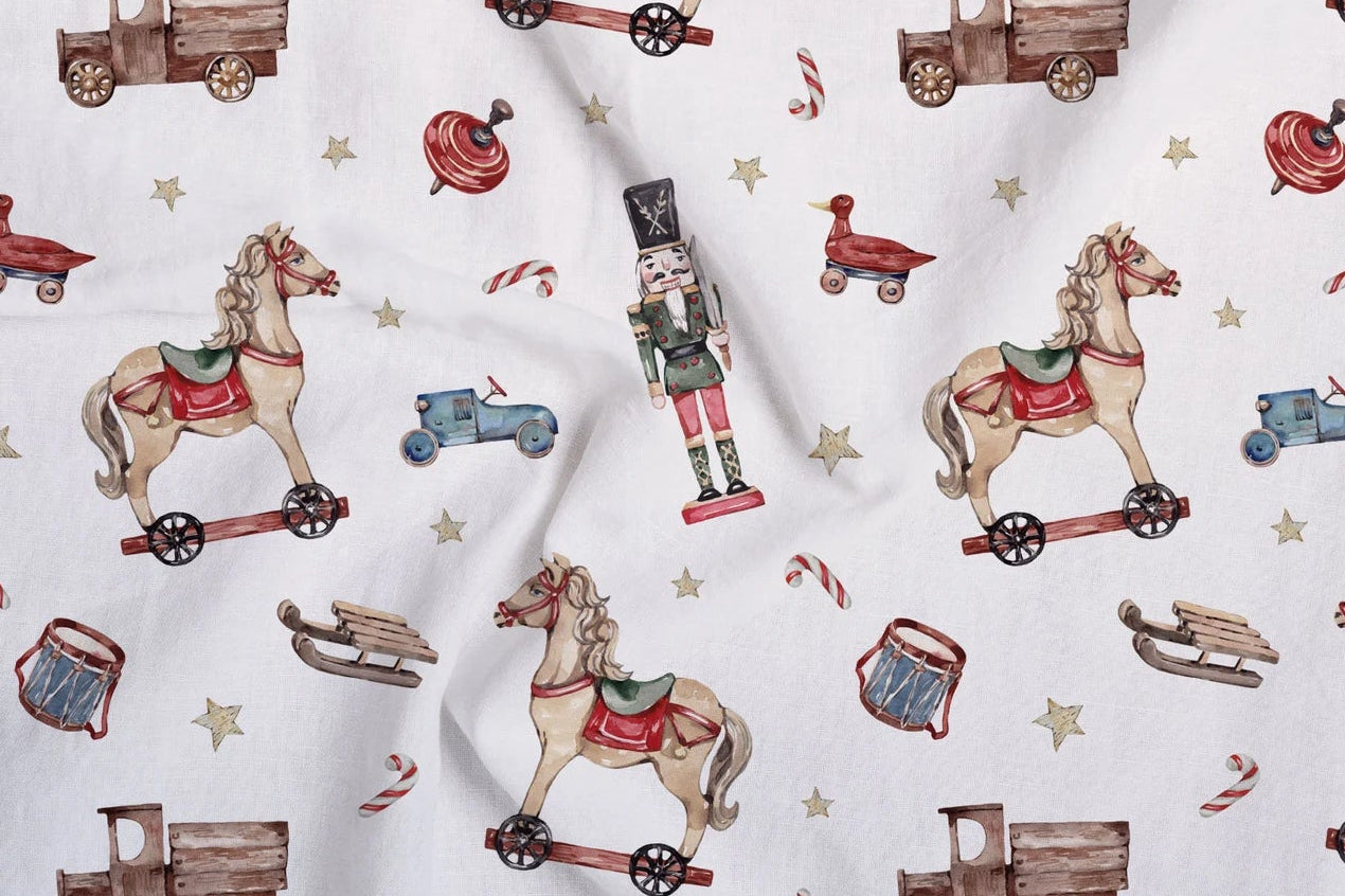 White fabric with a repeating pattern of vintage toy motifs including rocking horses on wheeled bases, nutcrackers, toy trucks, candy canes, sledding, and stars.