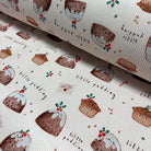 A close-up of a ribbed fabric or wallpaper with a repeating pattern of Christmas puddings, text reading 'little pudding', and small festive illustrations on an off-white background.