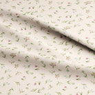 A close-up of draped fabric with a delicate all-over floral print on a warm cream background. The pattern features small green leaves, pink blossoms, and light blue flowers.