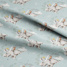 A close-up of a light blue fabric with a repeating pattern of white storks carrying baskets and the phrase 'little one' in script, with soft folds showing the material's texture.