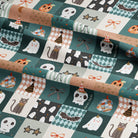 A close-up of a folded fabric with a repeating Halloween-themed patchwork print. The pattern features squares with ghosts, cats, skulls, bats, pumpkins, stars, and candy motifs in teal, beige, white, black, and orange tones.