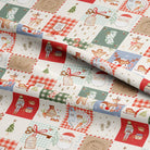 A close-up of a holiday-themed fabric with a patchwork design featuring Santa Claus, reindeer, nutcrackers, Christmas trees, gingham checks, and gift boxes in red, green, beige, and blue tones.