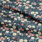 A close-up of a draped fabric with a dense floral print featuring white daisies, pink and peach flowers, and green leaves on a dark teal background.