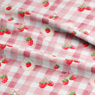 Pink and white gingham check fabric with a repeating pattern of red strawberries, green leaves, and small pink bows, shown folded to display the material and print.