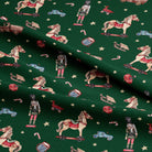 A close-up of dark green fabric printed with a holiday-themed pattern featuring toy horses, nutcrackers, candy canes, sleighs, and small vehicles, arranged in soft folds.