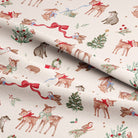 A close-up of cream-colored fabric with a festive Christmas print featuring brown deer, red bows, green holly, birds, and small holiday motifs, with the fabric folded diagonally.